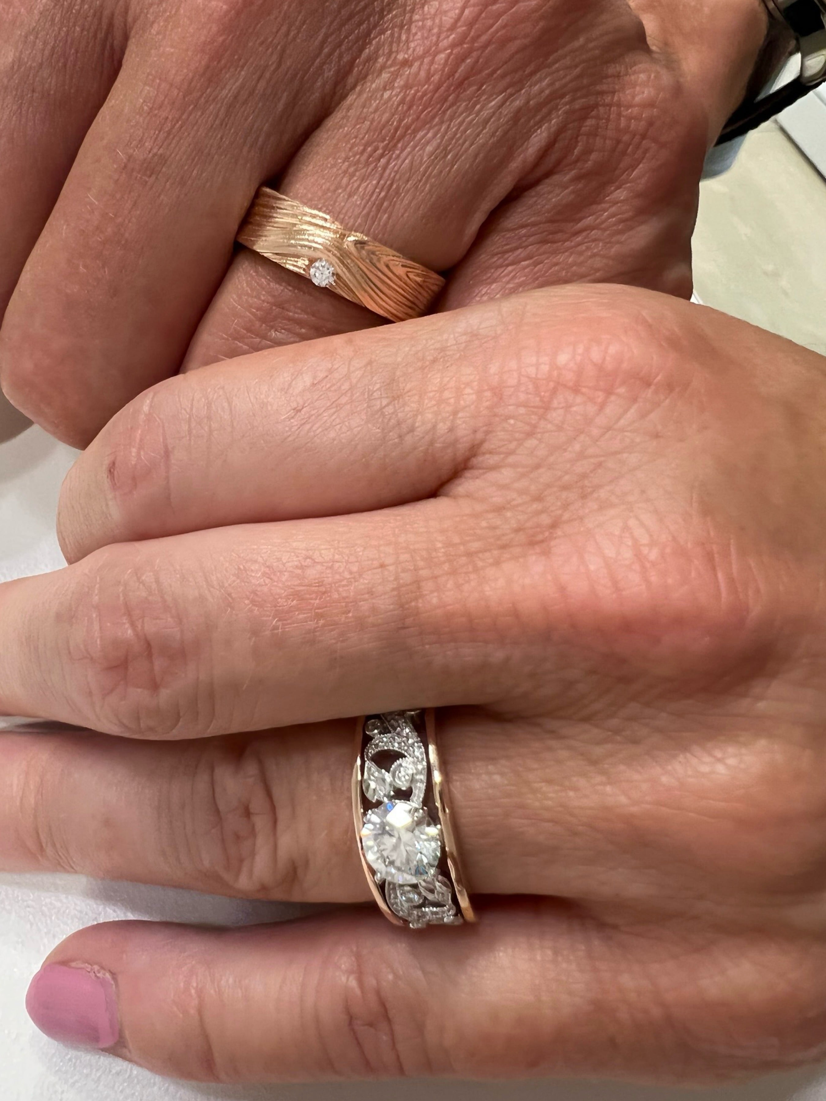 A 14k rose gold Barn Board wedding band displayed on a man’s hand with an 18k rose and white 1ct Enchanted Garden Engagement Ring on a woman’s hand.