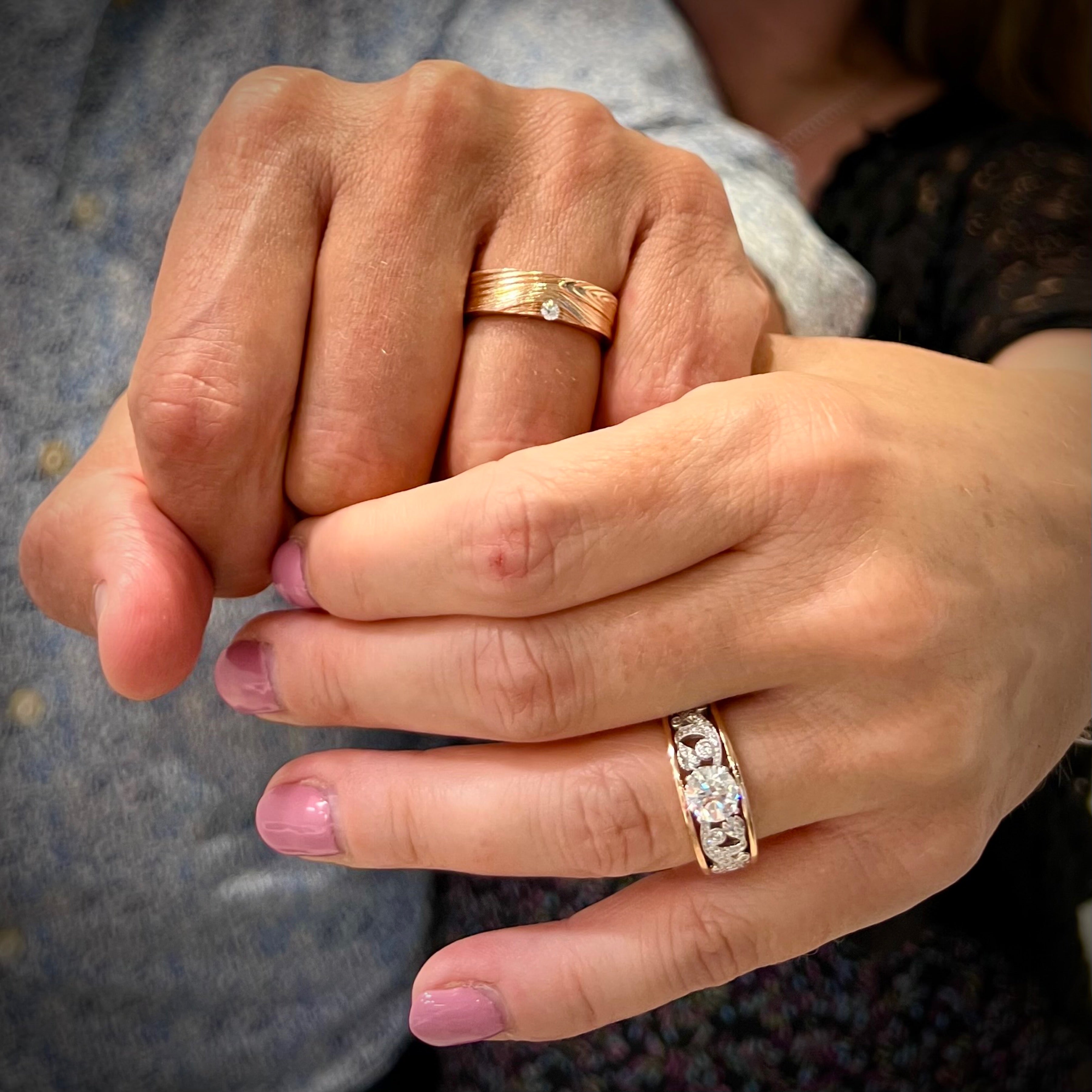 A 14k rose gold Barn Board wedding band with 0.06ct Canadian diamond displayed on a man’s hand with an Enchanted Garden Engagement Ring on a woman’s hand.