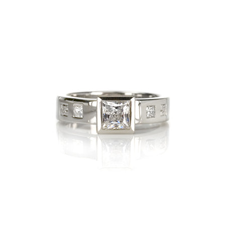 Crafted in 14KT white gold, this ring features a raised bezel set princess cut diamond. Band is set with princess-cut diamonds.