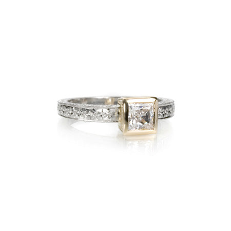 Crafted in 14KT white gold, this ring features a princess cut diamond set in a 14KT yellow gold square bezel setting on an orange blossom hand engraved band. 