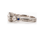 An ornate silver-toned ring featuring a large round clear gemstone, a smaller round clear gemstone, and a small round blue sapphire accent, set on a white background.