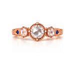 A rose gold three-stone ring featuring a large round center stone and two smaller round side stones, accented with tiny blue gemstones on the band, set against a clean white background.