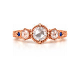 A rose gold three-stone ring featuring a large round center stone and two smaller round side stones, accented with tiny blue gemstones on the band, set against a clean white background.
