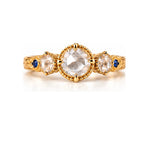 A yellow gold three-stone ring featuring a large oval clear gemstone in the center, flanked by two smaller oval clear gemstones, with small round blue accent gems on the band, set against a plain white background.