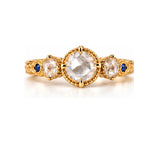 A yellow gold three-stone ring featuring a large oval clear gemstone in the center, flanked by two smaller oval clear gemstones, with small round blue accent gems on the band, set against a plain white background.