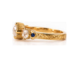 An ornate yellow gold ring featuring three diamonds in claw settings, a small round blue sapphire accent, and an engraved band, shown on a clean white background.