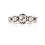 An antique-style three-stone ring featuring a large round-cut clear center stone, flanked by two smaller round-cut stones, with small blue accent stones on a silver-toned band, set against a plain white background.