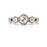 An antique-style three-stone ring featuring a large round-cut clear center stone, flanked by two smaller round-cut stones, with small blue accent stones on a silver-toned band, set against a plain white background.