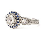 An ornate silver-toned engagement ring featuring a large round clear center stone surrounded by a halo of small dark blue stones, with a secondary clear stone and engraved detailing on the band, shown on a white background. Made to stack with collection rings