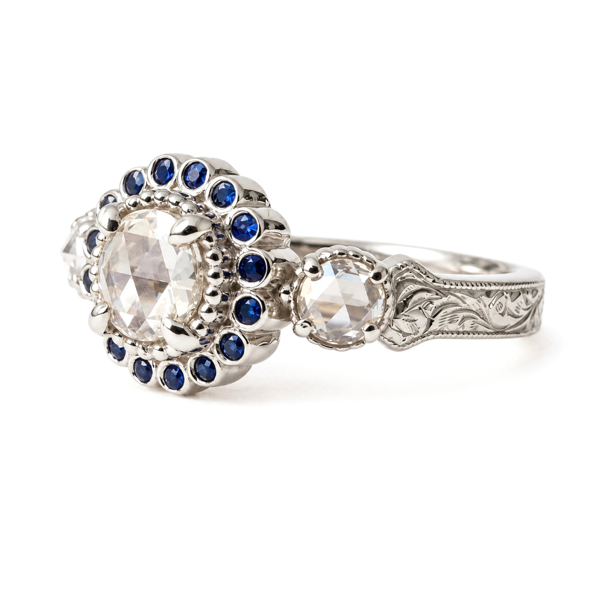 An ornate silver-toned engagement ring featuring a large round clear center stone surrounded by a halo of small dark blue stones, with a secondary clear stone and engraved detailing on the band, shown on a white background. Made to stack with collection rings
