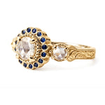 Crafted in 14KT yellow gold, this ring features a white rose-cut centre diamond with a blue sapphire halo and 2 smaller rose-cut diamonds on its sides.  All on a vintage-inspired hand engraved band. Can be an engagement ring or milestone ring.
