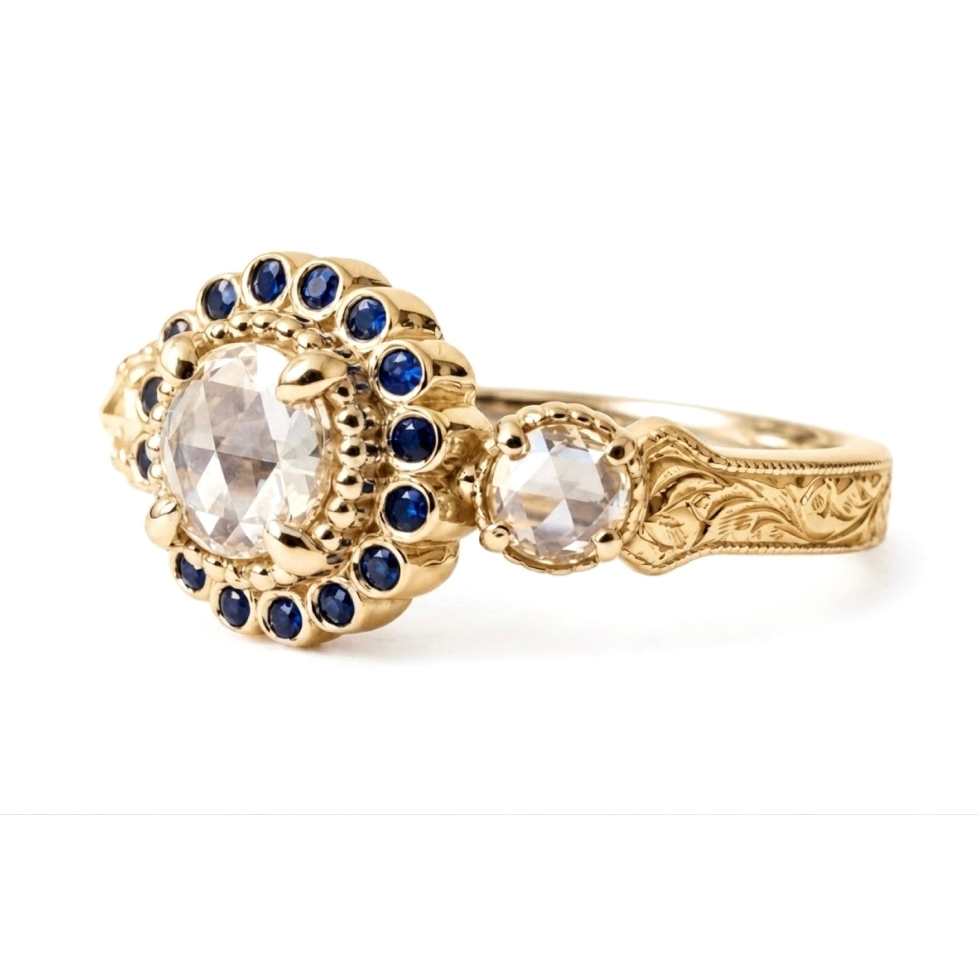 Crafted in 14KT yellow gold, this ring features a white rose-cut centre diamond with a blue sapphire halo and 2 smaller rose-cut diamonds on its sides.  All on a vintage-inspired hand engraved band. Can be an engagement ring or milestone ring.