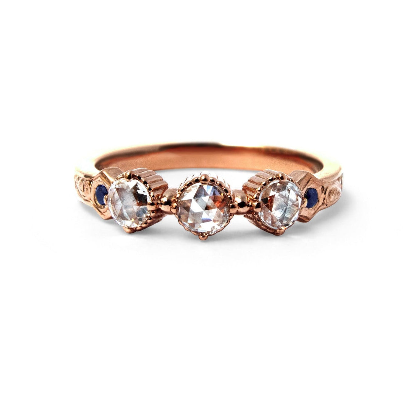 Crafted in 14KT rose gold, this ring features 3 round brilliant-cut diamonds in a row, with a blue sapphire on either side on a vintage-inspired hand engraved band.