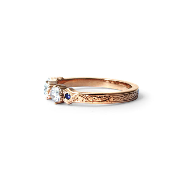 Crafted in 14KT rose gold, this ring features 3 round brilliant-cut diamonds in a row, with a blue sapphire on either side on a vintage-inspired hand engraved band.