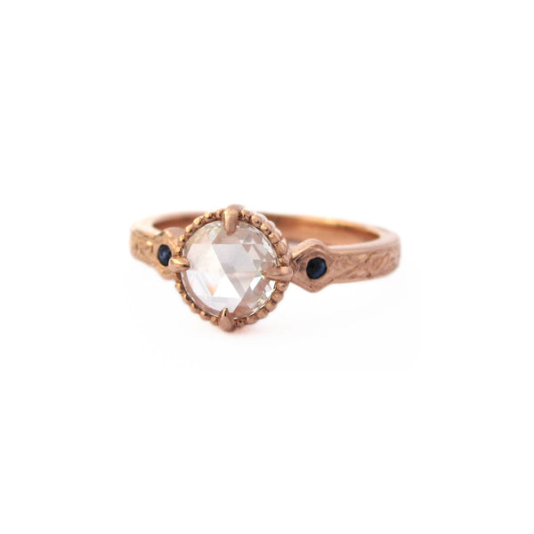 Crafted in 14KT rose gold, this ring features a large rose-cut diamond with two blue sapphires at its side on a vintage-inspired hand engraved band.