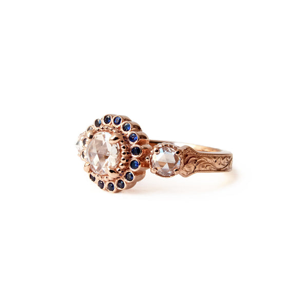 Crafted in 14KT rose gold, this ring features a white rose-cut centre diamond with a blue sapphire halo and 2 smaller rose-cut diamonds on its sides.  All on a vintage-inspired hand engraved band.