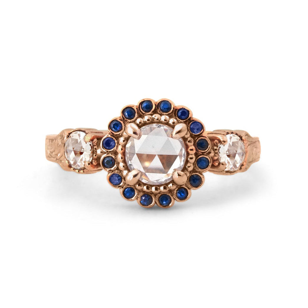 Crafted in 14KT rose gold, this ring features a white rose-cut centre diamond with a blue sapphire halo and 2 smaller rose-cut diamonds on its sides.  All on a vintage-inspired hand engraved band.