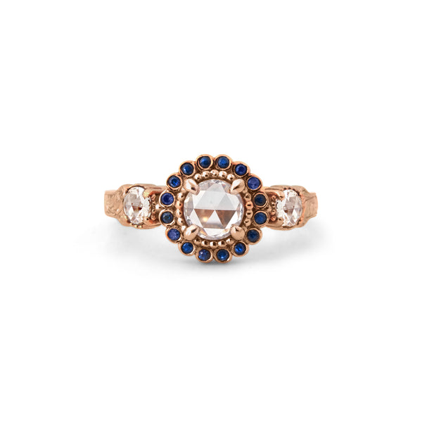 Crafted in 14KT rose gold, this ring features a white rose-cut centre diamond with a blue sapphire halo and 2 smaller rose-cut diamonds on its sides.  All on a vintage-inspired hand engraved band.