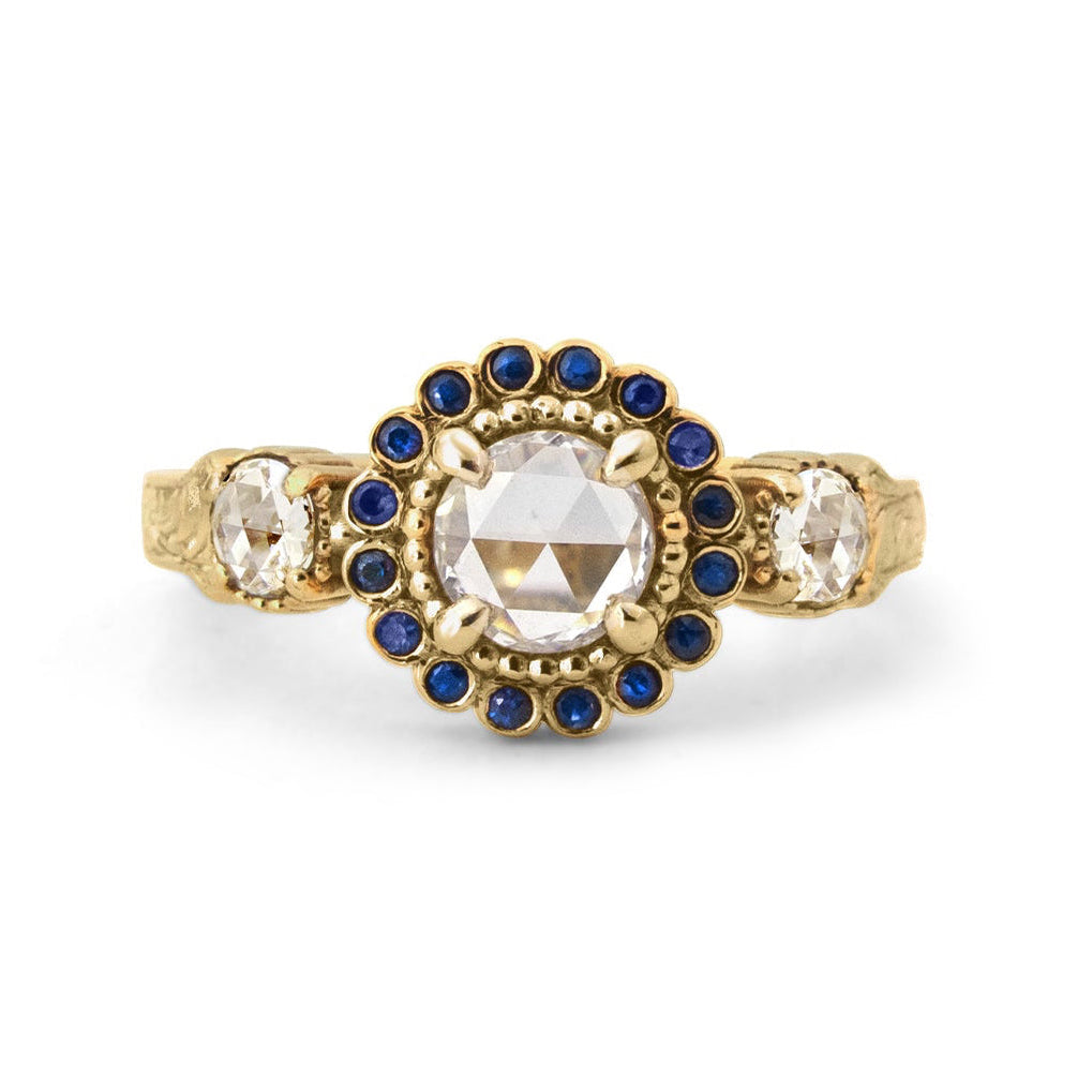 Crafted in 14KT yellow gold, this ring features a white rose-cut centre diamond with a blue sapphire halo and 2 smaller rose-cut diamonds on its sides.  All on a vintage-inspired hand engraved band.