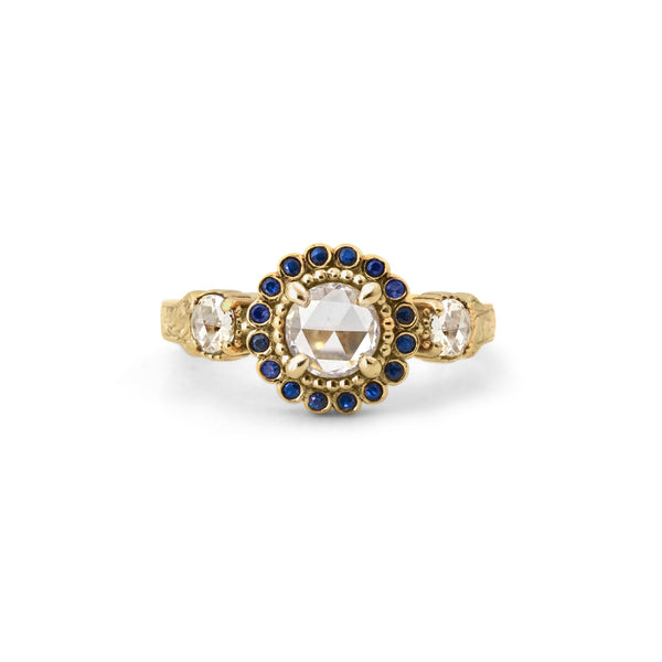 Crafted in 14KT yellow gold, this ring features a white rose-cut centre diamond with a blue sapphire halo and 2 smaller rose-cut diamonds on its sides.  All on a vintage-inspired hand engraved band.