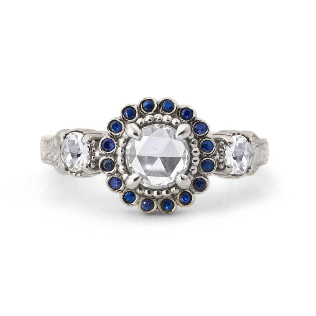 Crafted in 14KT white gold, this ring features a white rose-cut centre diamond with a blue sapphire halo and 2 smaller rose-cut diamonds on its sides.  All on a vintage-inspired hand engraved band.
