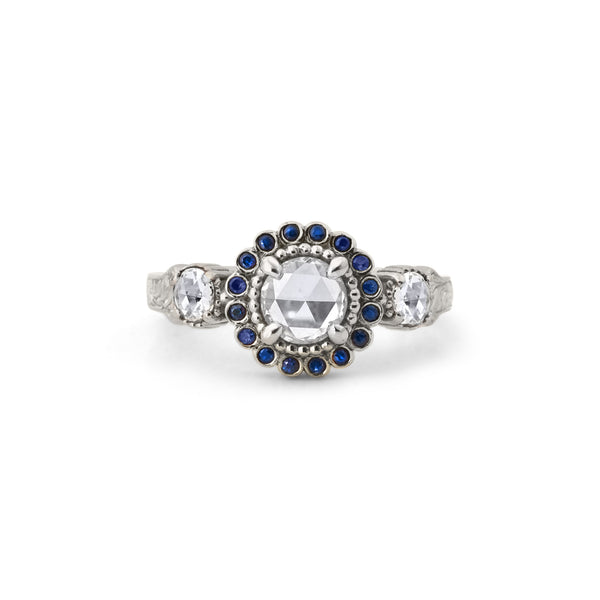 Crafted in 14KT white gold, this ring features a white rose-cut centre diamond with a blue sapphire halo and 2 smaller rose-cut diamonds on its sides.  All on a vintage-inspired hand engraved band.