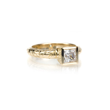 Crafted in 14KT yellow gold, this ring features a bezel set princess-cut diamond with accent diamonds on either side on a paisley hand-engraved band. 