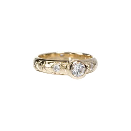 Crafted in 14KT yellow gold, this ring features a bezel set round brilliant-cut diamond with diamond accents on either side on a paisley hand engraved band.