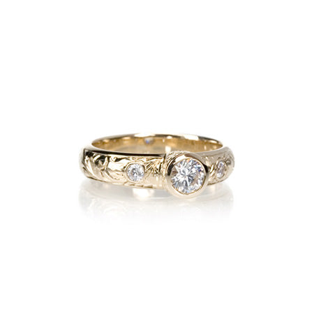 Crafted in 14KT yellow gold, this ring features a bezel set round brilliant-cut diamond with diamond accents on either side on a paisley hand engraved band.