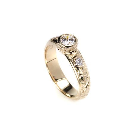 Crafted in 14KT yellow gold, this ring features a bezel set round brilliant-cut diamond with diamond accents on either side on a paisley hand engraved band.