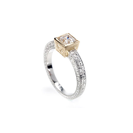Crafted in 14KT white gold, this ring features a princess cut diamond set in a 14KT yellow gold square bezel setting on an orange blossom hand engraved band. 